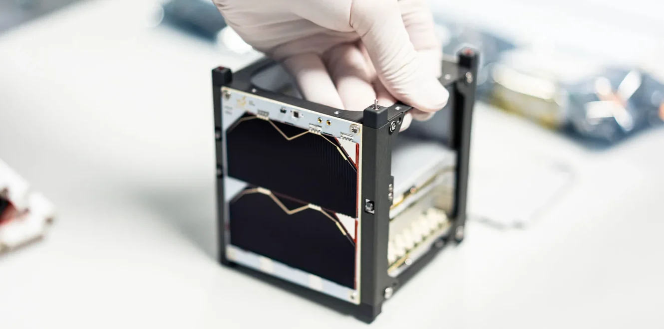 A top side view of the EnduroSat 1U CubeSat satellite bus in a cleanroom production facility. A gloved hand is holding top right edge of the 1U CubeSat structure as the nanosatellite is being constructed.