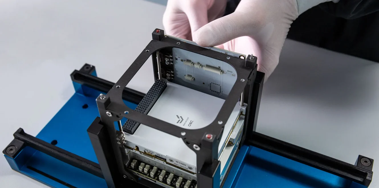 A top side view of the EnduroSat 1U CubeSat satellite bus in a cleanroom production facility as two hands are attaching the 1U Solar Panels X/Y on its side.